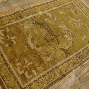 A distressed olive and brown traditional rug with geometric borders sits on rustic wooden floorboards.