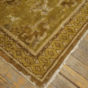 Worn, low-pile rug in olive green and brown with a geometric border on rustic floorboards.
