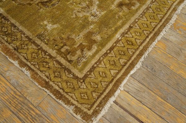 Worn, low-pile rug in olive green and brown with a geometric border on rustic floorboards.