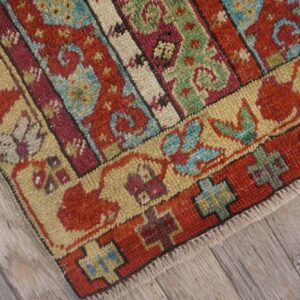 Knotted textile close-up shows red, green, and beige banded patterns resting on weathered grey wood floor.