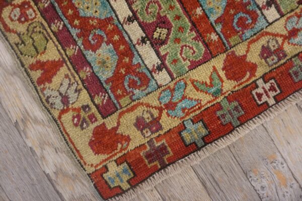 Knotted textile close-up shows red, green, and beige banded patterns resting on weathered grey wood floor.
