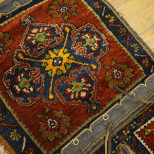 Rustic red and blue geometric rug attached to dark leather, resting on light wooden planks.