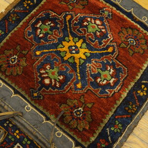 Knotted textile fragment in rust, navy, and gold patterns, edged with dark leather on plank wood flooring.