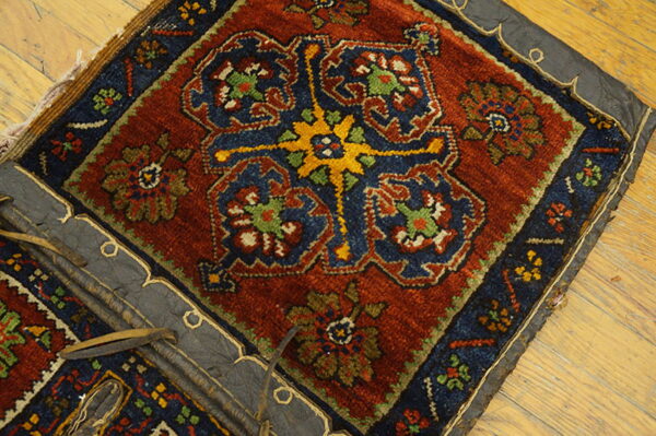 Knotted textile fragment in rust, navy, and gold patterns, edged with dark leather on plank wood flooring.