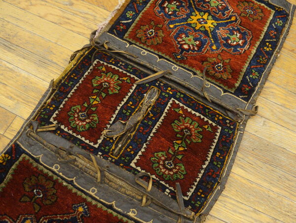 A detailed red and navy rug textile with floral medallions and dark leather straps rests on light wood floorboards.