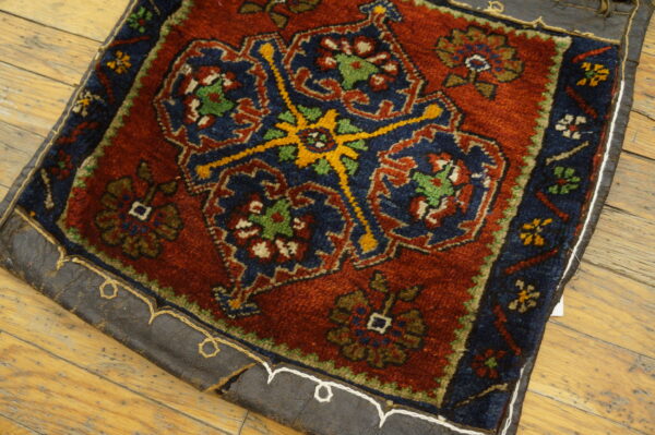 High-pile red and navy rug with geometric motifs and a leather stitched backing rests on distressed wood flooring.