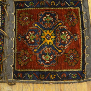 Rust and blue patterned textile fragment with leather trim and straps resting on light wood flooring.
