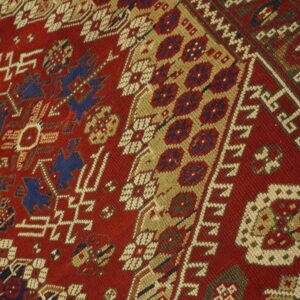Geometric patterns in red, tan, navy, and cream dominate this low-pile rug lying on a wood floor.