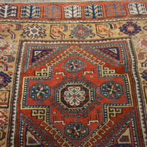 Red and navy geometric rug with a tan border sits on a weathered light brown wooden floor.