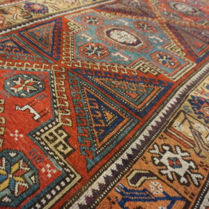 Geometric patterned rug with a main field of terracotta red, blue, and teal on wooden flooring.