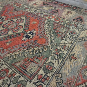 Low-pile geometric rug in distressed red, green, and beige, displayed on light gray wood flooring.