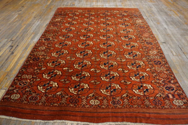 Rust-colored rug featuring navy and cream repeating geometric gul motifs on a distressed wood floor.