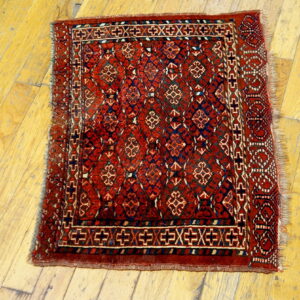 Red geometric rug with dark blue motifs and fringed edges lies on worn yellow hardwood.
