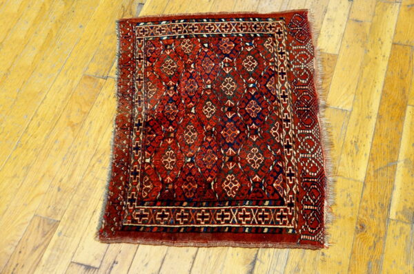 Red geometric rug with dark blue motifs and fringed edges lies on worn yellow hardwood.
