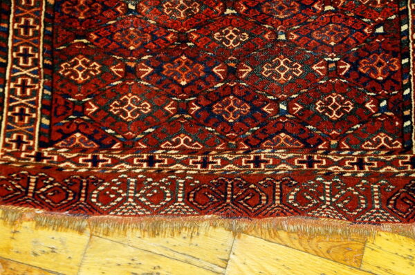 Red and navy geometric patterned rug fringe laying on light golden hardwood planks.