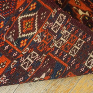 Geometric pattern rug corner folded over, displaying red, burgundy, navy, and white on wood flooring.