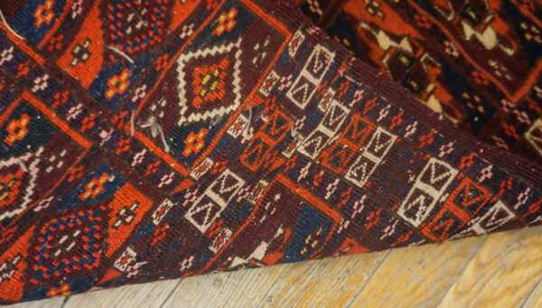 Geometric pattern rug corner folded over, displaying red, burgundy, navy, and white on wood flooring.