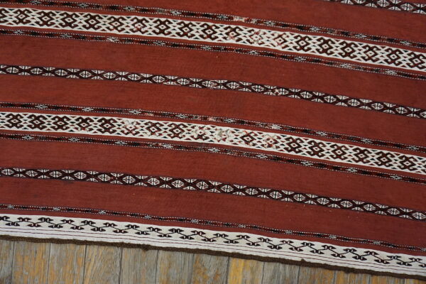 Terracotta flatweave rug featuring intricate white and dark brown geometric stripes over weathered wood flooring.