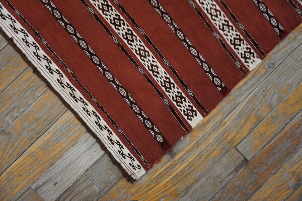 Red-brown, white, and dark striped flatweave rug displaying geometric patterns rests on rustic wooden planks.