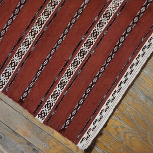 Flat-woven rug displaying rusty red and geometric white stripes sits on distressed light wood floorboards.