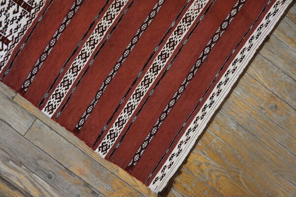 Flat-woven rug displaying rusty red and geometric white stripes sits on distressed light wood floorboards.