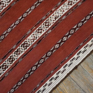Rust flatweave rug with white and dark geometric striped patterns lies diagonally on weathered wood planks.