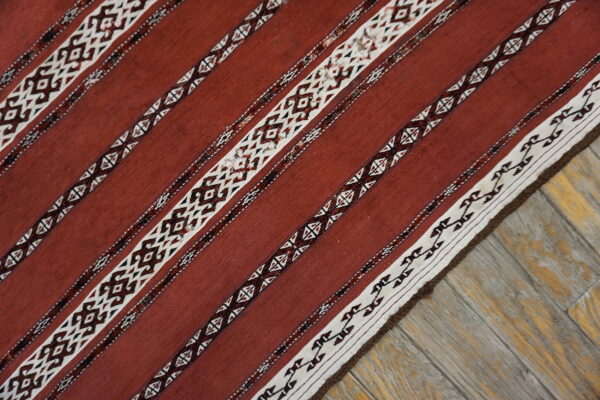 Rust flatweave rug with white and dark geometric striped patterns lies diagonally on weathered wood planks.