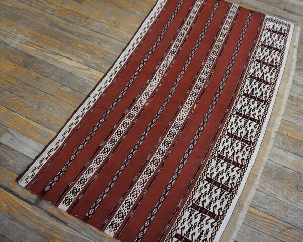 Rust-colored flatweave rug with white and dark brown geometric stripes displayed on weathered wooden planks.