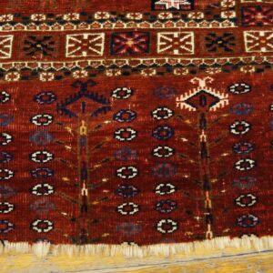 Close view of a red rug with geometric borders, repeating medallions, and fringe on yellow wood floor.