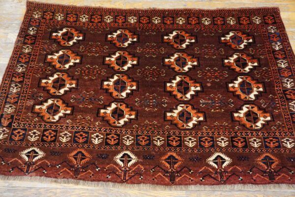 Dark red-brown high-pile rug displays repeating geometric medallions and detailed borders on light wood floor.