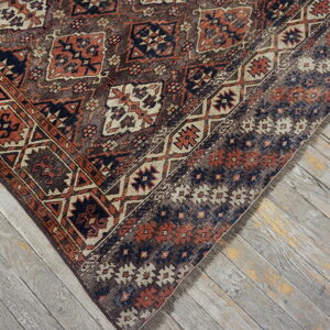 A low-pile rug corner displaying geometric rust and dark blue patterns against distressed grey wood flooring.