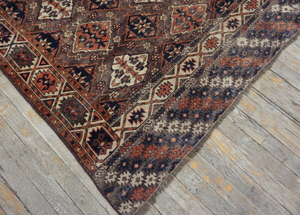 A low-pile rug corner displaying geometric rust and dark blue patterns against distressed grey wood flooring.