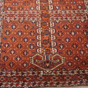Low-pile red rug with dense geometric patterns in blue, orange, and cream on light-colored flooring.
