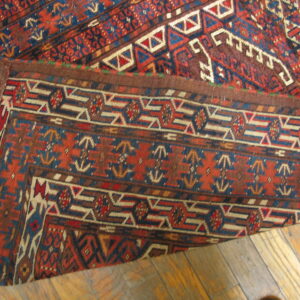 A richly colored rug with geometric red, blue, and brown patterns is folded over on a plank wood floor.