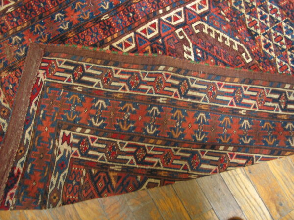 A richly colored rug with geometric red, blue, and brown patterns is folded over on a plank wood floor.