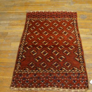 Reddish-orange rug with geometric patterns in navy and cream on scratched wood flooring.