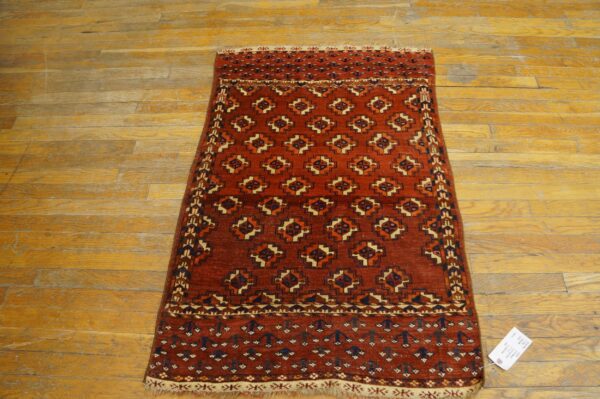 Reddish-orange rug with geometric patterns in navy and cream on scratched wood flooring.