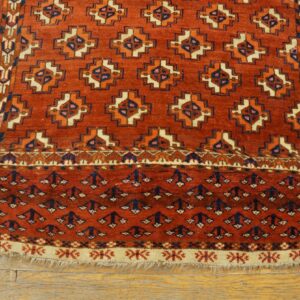 A red-orange geometric rug with navy, cream, and orange designs lying on light wood planks.