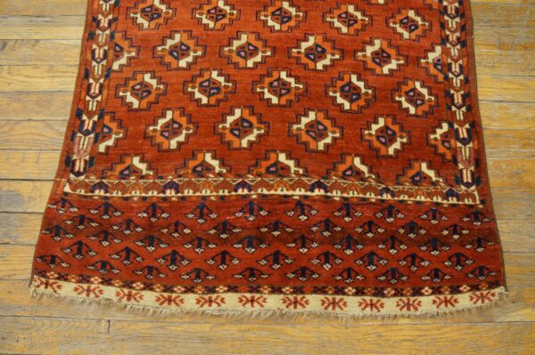 A red-orange geometric rug with navy, cream, and orange designs lying on light wood planks.