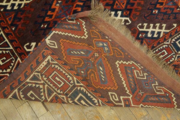 Geometric brown, red, and blue rug folded back, exposing its kilim end and fringe on a wood floor.
