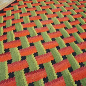 Low-profile rug featuring interlocking geometric patterns in bright red, olive green, and black on wood flooring.