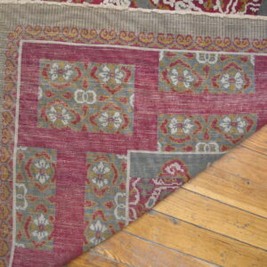 A red, gray, and gold patterned rug corner is folded back, revealing its woven reverse on a wood floor.