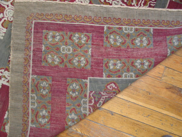 A red, gray, and gold patterned rug corner is folded back, revealing its woven reverse on a wood floor.