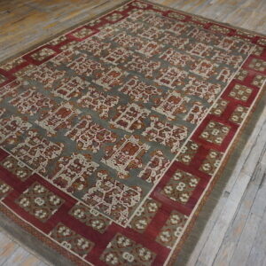 Muted gray and red rug with stylized pattern lies on distressed, wide-plank wood flooring.