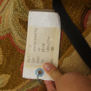 Close-up of a textural hooked rug in brown, green, and pink, partially covered by a label and black strap.