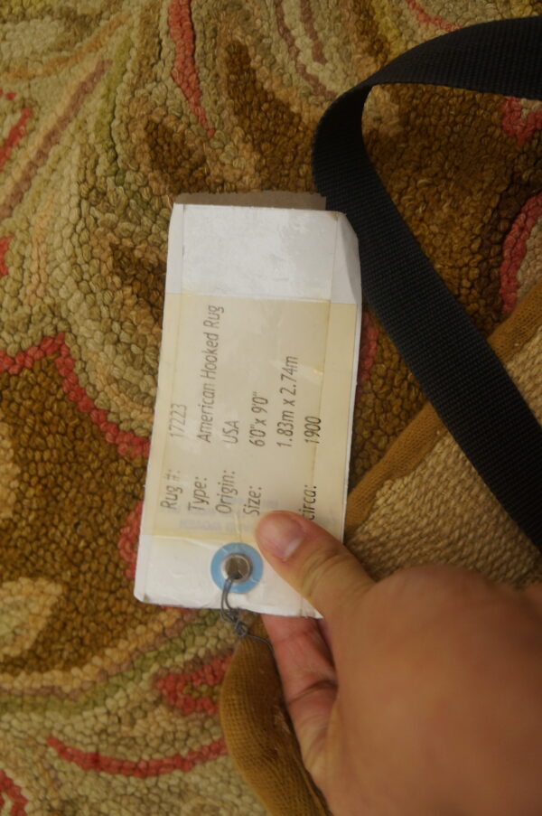 Close-up of a textural hooked rug in brown, green, and pink, partially covered by a label and black strap.