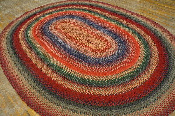 Multicolored braided oval rug featuring concentric bands of red, blue, green, and gray on a wood floor.