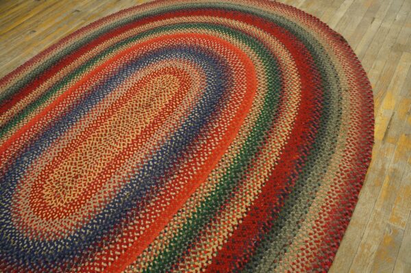A textured, braided, multicolored oval rug with banded rings lies on light, worn wooden flooring.