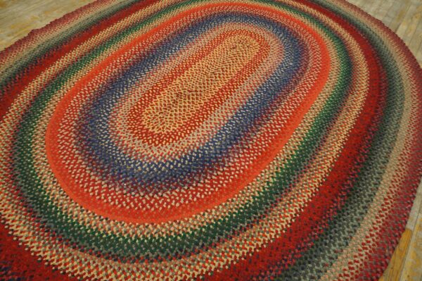 An oval braided rug with thick concentric stripes of red, blue, green, and gray upon wood flooring.