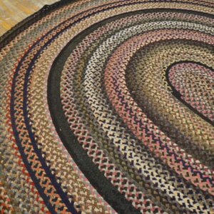 Round, braided rug displaying variegated rings of pink, brown, navy, and rust resting on aged wooden floorboards.
